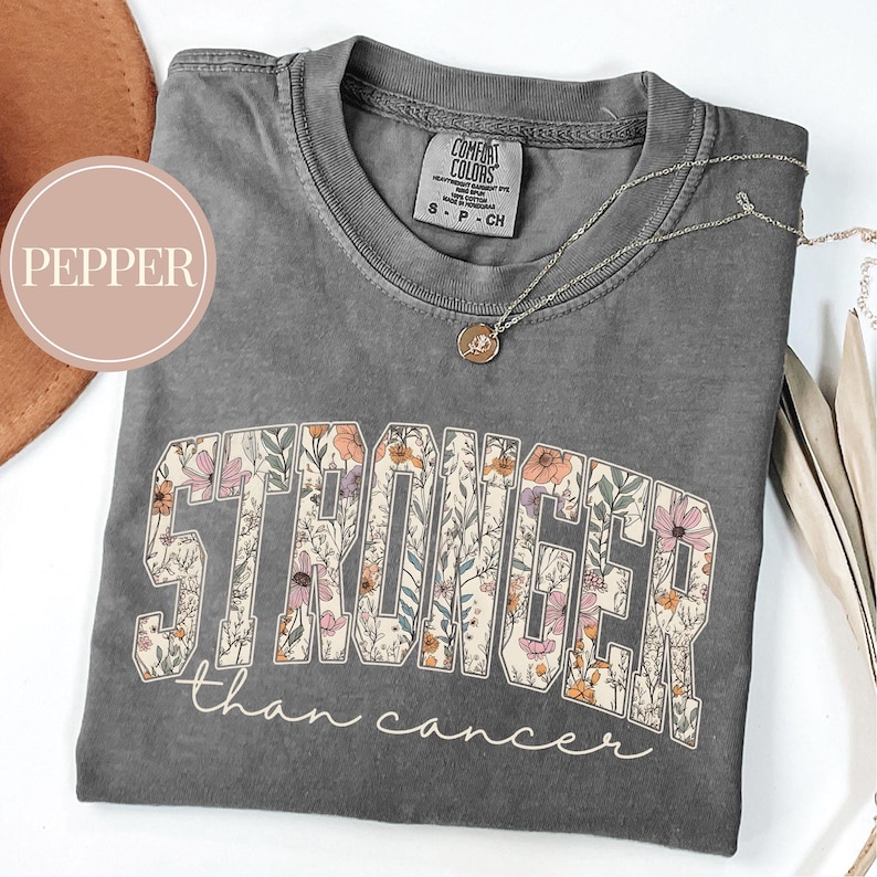 May include: A gray Comfort Colors t-shirt with the words "STRONGER than cancer" in a floral design. A silver necklace with a small pendant rests on the shirt. The shirt is folded.