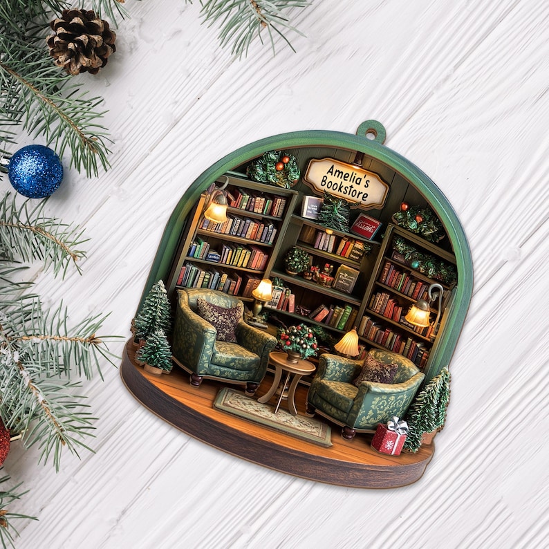 May include: A semi-circular hanging ornament depicting a miniature bookstore interior with two green patterned armchairs, a small round table, and shelves filled with books. A sign above the shelves reads Amelia's Bookstore. Includes miniature trees and a gift box.