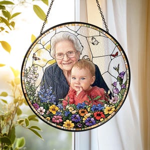 May include: Round stained glass window art featuring a portrait of a smiling elderly woman and a baby. The artwork includes butterflies and colorful flowers. The window is framed in black and hangs from a chain.