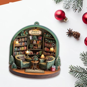 May include: A semi-circular hanging ornament depicting a miniature bookstore interior with two green patterned armchairs, a small round table, and shelves filled with books. A sign reads Amelia's Bookstore. The scene includes warm lighting and small decorative pine trees.