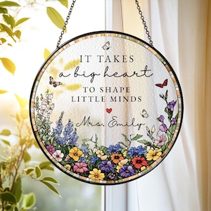 May include: A circular stained glass suncatcher with a black metal frame and hanging chain. The glass features a floral border with colourful flowers and butterflies, and the text: IT TAKES a big heart TO SHAPE LITTLE MINDS, with a red heart and Mrs. Emily.