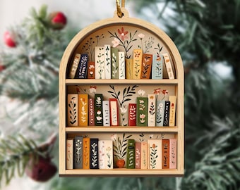 May include: Wooden bookshelf ornament with a gold string for hanging. The ornament features a miniature bookshelf design with painted books in various colors and floral accents. The background is a blurred Christmas tree.