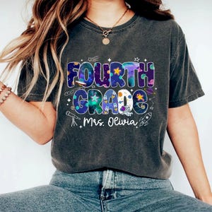 May include: Dark gray t-shirt with a colorful "FOURTH GRADE" graphic. The design features a space theme with stars, planets, and a rocket, along with the name "Mrs. Olivia". A casual, comfortable tee for students or teachers.