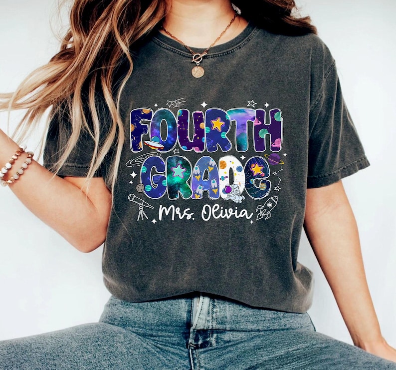 May include: Dark gray t-shirt with a colorful "FOURTH GRADE" graphic. The design features a space theme with stars, planets, and a rocket, along with the name "Mrs. Olivia". A casual, comfortable tee for students or teachers.