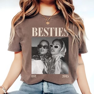 May include: A brown t-shirt featuring a black and white image of two people wearing sunglasses and headscarves. The word "BESTIES" is printed above the image, with "EST. 2015" below. The shirt has short sleeves.