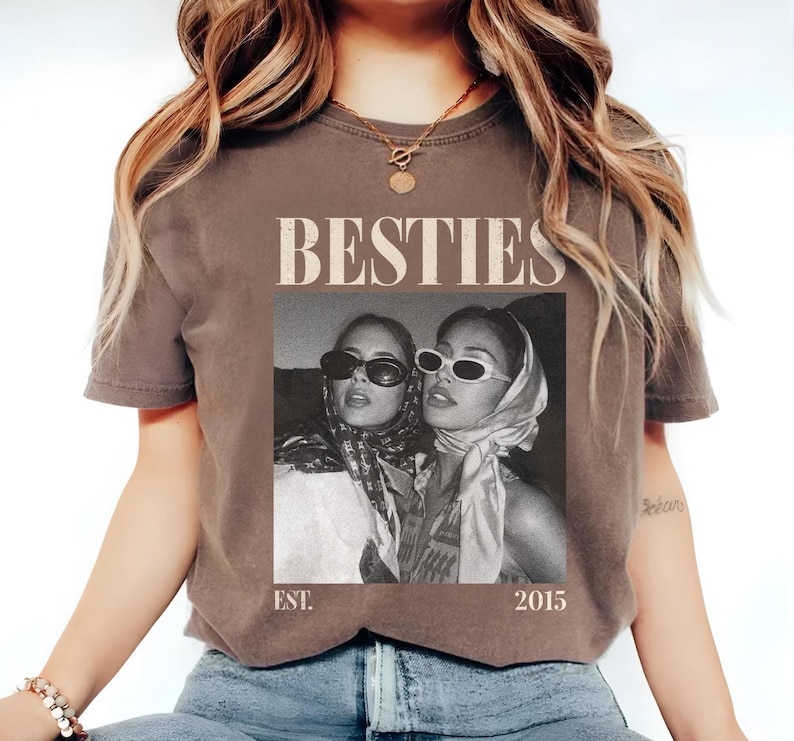 May include: A brown t-shirt featuring a black and white image of two people wearing sunglasses and headscarves. The word "BESTIES" is printed above the image, with "EST. 2015" below. The shirt has short sleeves.