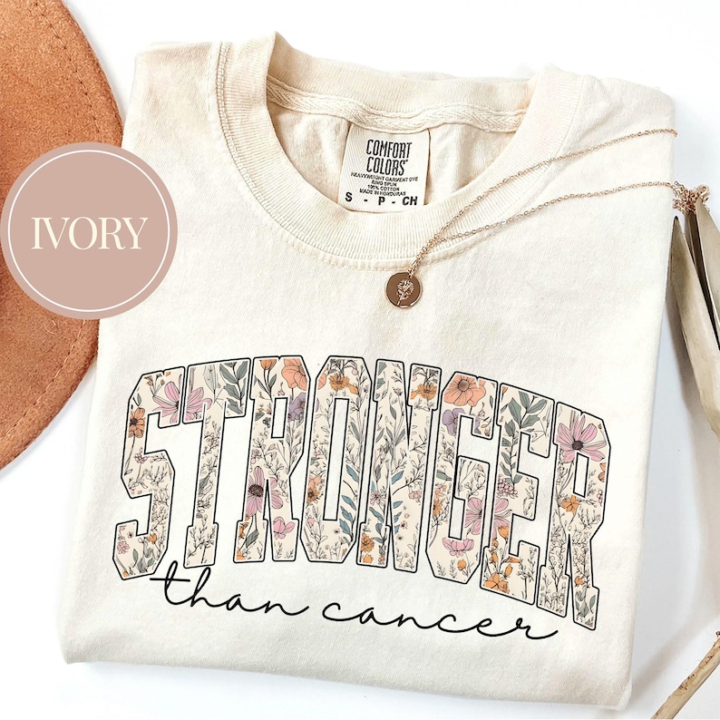 May include: Ivory-colored t-shirt with the words "STRONGER" and "than cancer" in a floral design. The shirt has a crew neck and short sleeves. A gold necklace with a pendant is draped on the shirt.