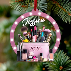 May include: A pink Christmas ornament with the name "Jessica" in cursive. The ornament features a ring of lights around the edge and a makeup bag filled with brushes, lipsticks, and nail polish. The year "2024" is printed on the bag.