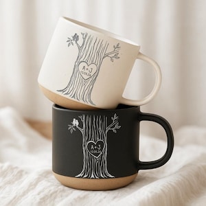 May include: Two ceramic mugs stacked vertically, one cream and one black. Each features a tree illustration with two birds perched on a branch and a heart carved into the trunk containing the text A + J and the date 10.08.26 in a white or grey ink.