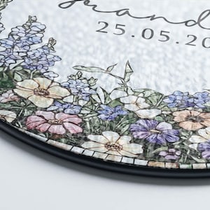 May include: Round decorative piece with a floral mosaic design. The mosaic features a variety of flowers in shades of white, pink, purple, and blue, with green foliage. The text "grand" and the date "25.05.20" are visible.
