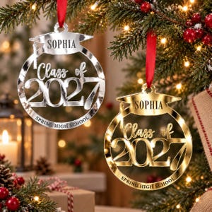 May include: Two circular graduation ornaments, one silver and one gold, each featuring a graduation cap at the top, the name SOPHIA, the text Class of 2027, and the words SPRING HIGH SCHOOL, all suspended by red ribbons from a pine tree branch.