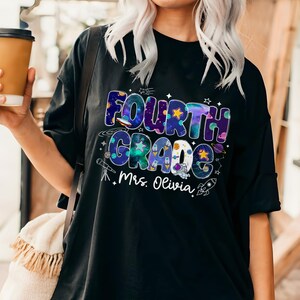 May include: Black t-shirt with a colorful Fourth Grade graphic. The design includes the words "Fourth Grade" in a space-themed font, along with stars, planets, and a rocket ship. The name "Mrs. Olivia" is printed below the graphic.