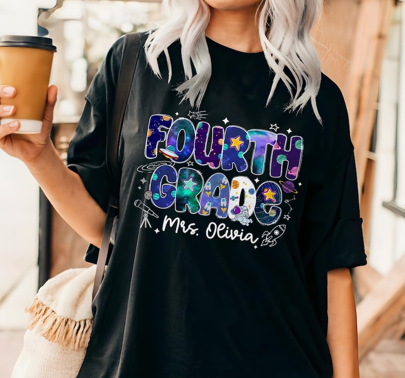 May include: Black t-shirt with a colorful Fourth Grade graphic. The design includes the words "Fourth Grade" in a space-themed font, along with stars, planets, and a rocket ship. The name "Mrs. Olivia" is printed below the graphic.