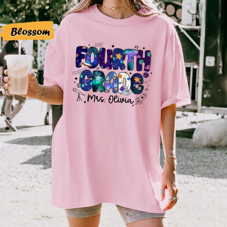 May include: A light pink Fourth Grade t-shirt with a colorful space-themed design. The design includes the words "Fourth Grade" in a bubble font, along with stars, planets, and a rocket ship. The text "Mrs. Olivia" is also included.