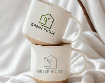 May include: Two off-white ceramic mugs stacked vertically. Each mug features a speckled texture, a smooth handle, and a raw clay base. The top mug displays a green house icon with the text GREEN HOUSE, while the bottom mug shows a grey version.