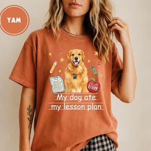 Puede incluir: Una camiseta de manga corta color &oacute;xido con un gr&aacute;fico de un perro golden retriever, un l&aacute;piz, un cray&oacute;n, un documento de plan de lecciones y una manzana roja. El texto "My dog ate my lesson plan" est&aacute; impreso en blanco bajo el perro.