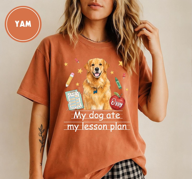 Puede incluir: Una camiseta de manga corta color &oacute;xido con un gr&aacute;fico de un perro golden retriever, un l&aacute;piz, un cray&oacute;n, un documento de plan de lecciones y una manzana roja. El texto "My dog ate my lesson plan" est&aacute; impreso en blanco bajo el perro.