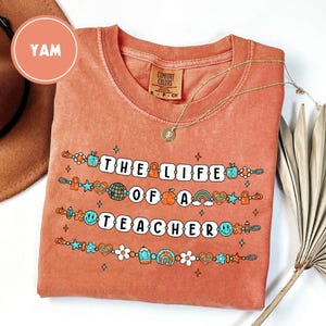 May include: A coral-colored t-shirt with the text "THE LIFE OF A TEACHER" in white block letters. The shirt features colorful graphics of apples, rainbows, and stars. A gold necklace with a pendant is draped on the shirt.