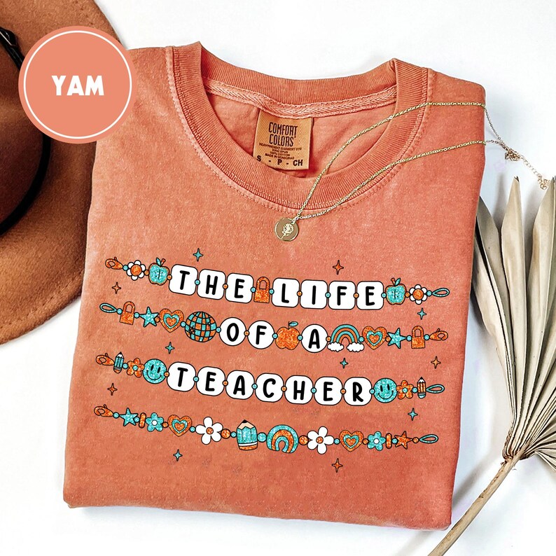 May include: A coral-colored t-shirt with the text "THE LIFE OF A TEACHER" in white block letters. The shirt features colorful graphics of apples, rainbows, and stars. A gold necklace with a pendant is draped on the shirt.