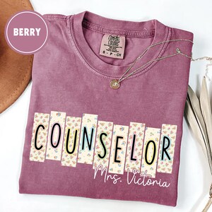 May include: Berry-colored t-shirt with the word "COUNSELOR" in a floral design, with "Mrs. Victoria" below. The shirt has a Comfort Colors tag and a silver necklace. A berry-colored circle is in the upper left corner.
