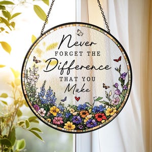 May include: A circular stained glass suncatcher featuring a black frame and a chain for hanging. The center displays the text "Never FORGET THE Difference THAT YOU Make" with a small red heart, surrounded by colorful flowers and butterflies.