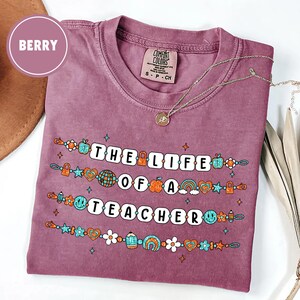 May include: Berry-colored t-shirt with the text "THE LIFE OF A TEACHER" in white block letters. The text is decorated with colorful illustrations of apples, rainbows, and pencils. A silver necklace with a pendant rests on the shirt.