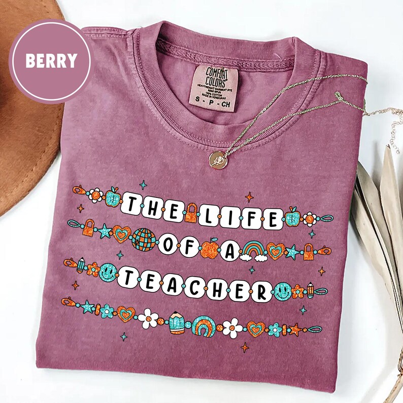 May include: Berry-colored t-shirt with the text "THE LIFE OF A TEACHER" in white block letters. The text is decorated with colorful illustrations of apples, rainbows, and pencils. A silver necklace with a pendant rests on the shirt.