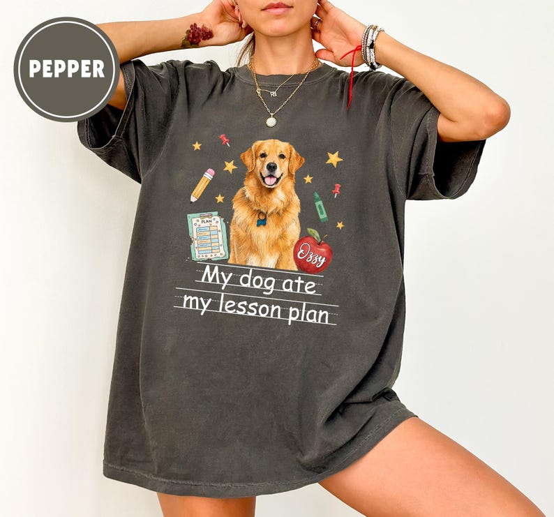 Puede incluir: Una camiseta gris carb&oacute;n de manga corta con un gr&aacute;fico de un perro golden retriever, una manzana roja con el nombre Ozzy, un l&aacute;piz y un documento de plan de lecci&oacute;n. El texto My dog ate my lesson plan est&aacute; impreso en blanco bajo el perro.