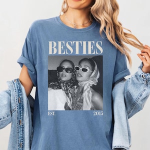 May include: A blue t-shirt with the word "BESTIES" in a bold, white font. Below the text is a black and white image of two women wearing sunglasses and headscarves. The shirt also has the text "EST. 2015".