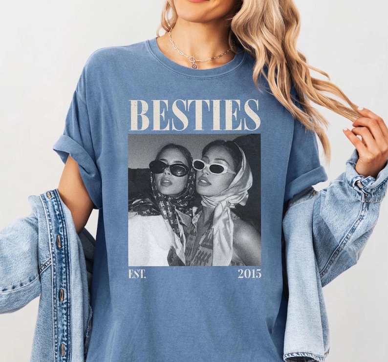 May include: A blue t-shirt with the word "BESTIES" in a bold, white font. Below the text is a black and white image of two women wearing sunglasses and headscarves. The shirt also has the text "EST. 2015".