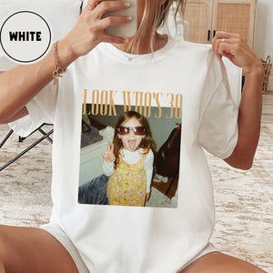 Może przedstawiać: Biała koszulka z napisem "LOOK WHO'S 30". Grafika przedstawia małą dziewczynkę w okularach przeciwsłonecznych, kt&oacute;ra pokazuje język. Dziewczynka ma na sobie ż&oacute;łtą sukienkę w kwiaty.
