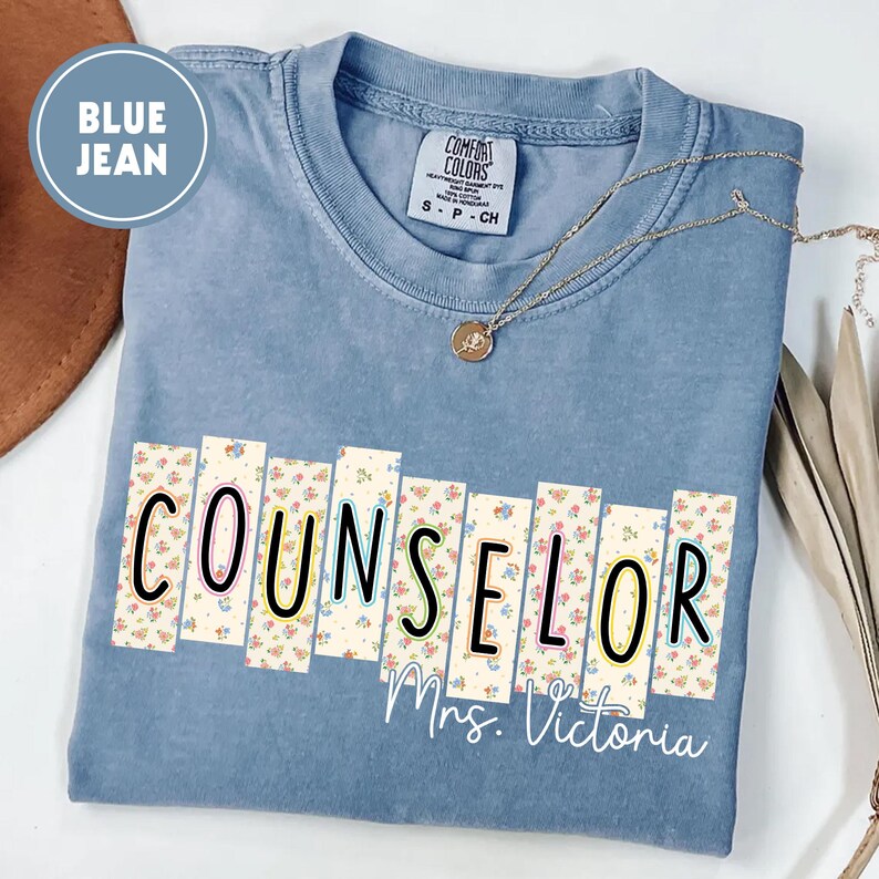 May include: A blue jean Comfort Colors t-shirt with the word "COUNSELOR" in a floral design. The name "Mrs. Victoria" is printed below the word. A gold necklace with a pendant is draped on the shirt.