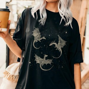 K&ouml;nnte beinhalten: Schwarzes T-Shirt mit drei detaillierten Drachenillustrationen mit ausgebreiteten Fl&uuml;geln, vor einem sternenklaren Nachthimmel. Die Drachen sind hellfarbig, was einen Kontrast zum dunklen Hintergrund erzeugt. Das Shirt hat eine lockere Passform.