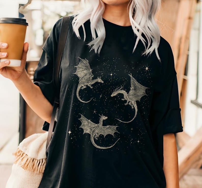 K&ouml;nnte beinhalten: Schwarzes T-Shirt mit drei detaillierten Drachenillustrationen mit ausgebreiteten Fl&uuml;geln, vor einem sternenklaren Nachthimmel. Die Drachen sind hellfarbig, was einen Kontrast zum dunklen Hintergrund erzeugt. Das Shirt hat eine lockere Passform.