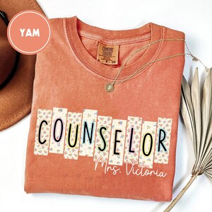 May include: A coral-colored t-shirt with the word "COUNSELOR" in a floral design. The name "Mrs. Victoria" is printed below the word. A gold necklace with a pendant is draped on the shirt.