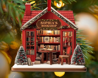 May include: A red Christmas ornament shaped like a miniature bookstore, with the text "SOPHIA'S BOOKSHOP" on a sign. The ornament features a detailed interior with bookshelves, a table, a chair, and small Christmas trees.