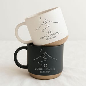 May include: Two stacked ceramic mugs, one cream and one black, each with a mountain design and the text "SOPHIA + DANIEL 10.20.2025". The mugs have a natural, unglazed base and a curved handle.