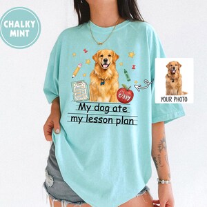 Puede incluir: Una camiseta azul claro con un gr&aacute;fico de un perro golden retriever, una manzana roja con el nombre Ossy, un l&aacute;piz, un cray&oacute;n y un documento de plan de lecci&oacute;n. El texto My dog ate my lesson plan est&aacute; impreso en negro bajo el perro.
