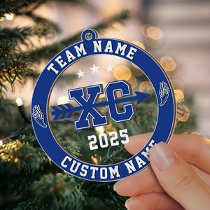 Puede incluir: Adorno navide&ntilde;o azul y blanco con el texto "TEAM NAME" y "CUSTOM NAME" en el borde. El centro presenta "XC" con una flecha, el a&ntilde;o "2025" y gr&aacute;ficos de alas. El adorno se sostiene en una mano.