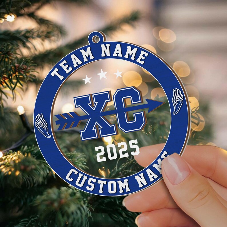 Puede incluir: Adorno navide&ntilde;o azul y blanco con el texto "TEAM NAME" y "CUSTOM NAME" en el borde. El centro presenta "XC" con una flecha, el a&ntilde;o "2025" y gr&aacute;ficos de alas. El adorno se sostiene en una mano.