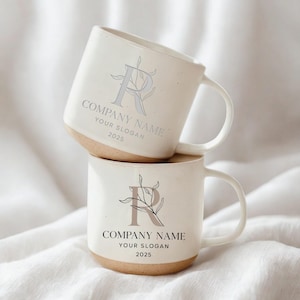 May include: Two stacked white ceramic mugs with a light brown base. Each mug features a stylized letter "R" with botanical accents and the text "COMPANY NAME YOUR SLOGAN 2025" in a light gray color. The mugs have a simple, elegant design.