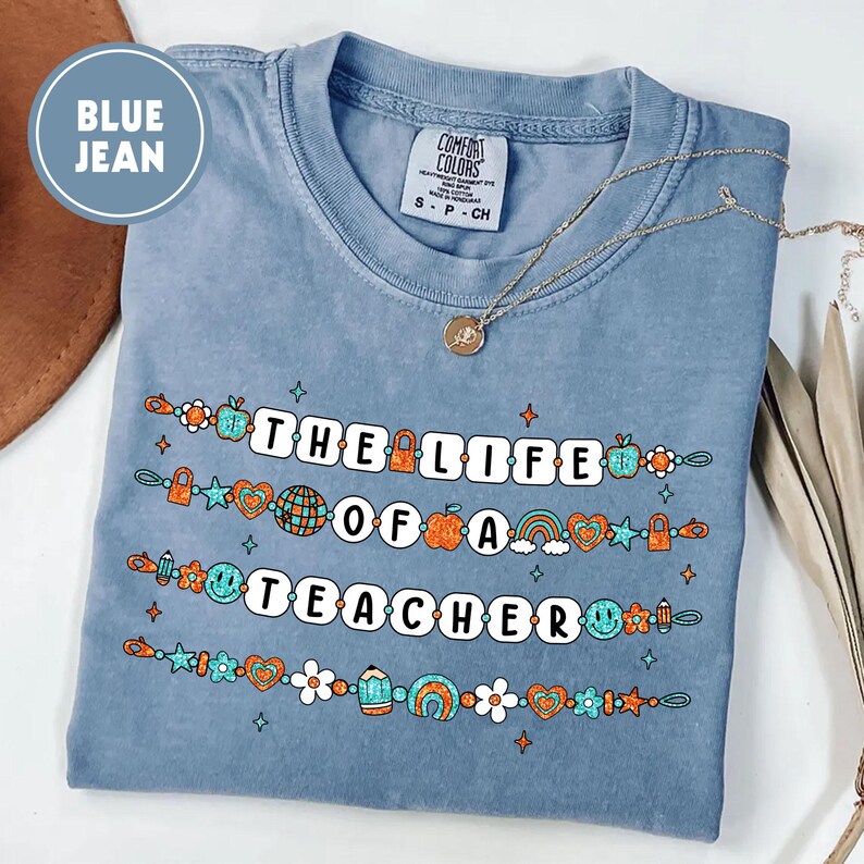 May include: Blue jean colored t-shirt with the text "THE LIFE OF A TEACHER" in white block letters. The text is decorated with colorful school-themed graphics. A gold necklace with a pendant rests on the shirt.