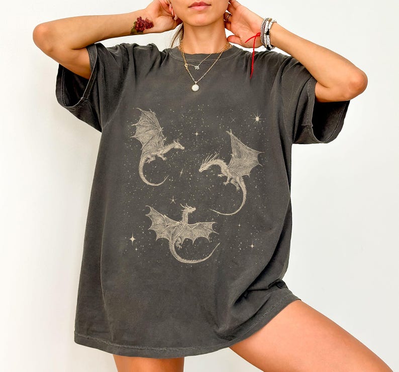 K&ouml;nnte beinhalten: Dunkelgraues T-Shirt mit einem Himmelsdesign mit drei Drachen und Sternen. Die Drachen sind im Vintage-Stil mit detaillierten Fl&uuml;geln und K&ouml;rpern dargestellt. Das T-Shirt hat eine lockere Passform und kurze &Auml;rmel.