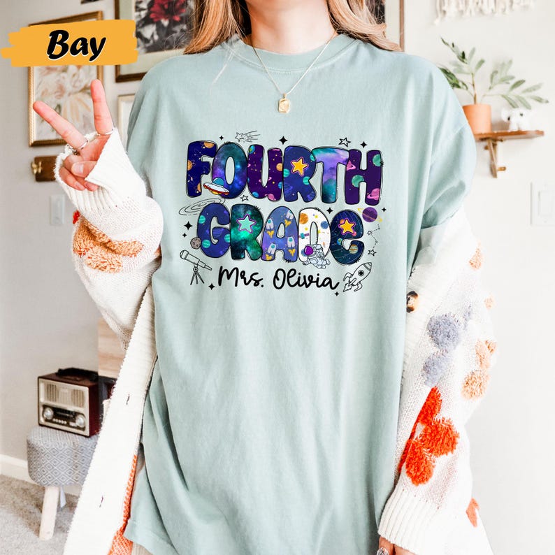 May include: A light blue t-shirt with the words "Fourth Grade" in a colorful space-themed design. The shirt also includes the name "Mrs. Olivia" and illustrations of a telescope and rocket ship.