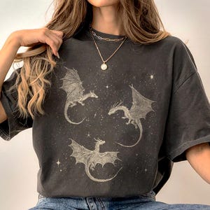K&ouml;nnte beinhalten: Dunkelgraues T-Shirt mit drei detaillierten Drachenillustrationen mit ausgebreiteten Fl&uuml;geln vor einem Sternenhintergrund. Die Drachen sind in einer hellen Farbe gehalten, die einen Kontrast zur Farbe des Shirts bildet. Das Shirt hat eine lockere Passform.