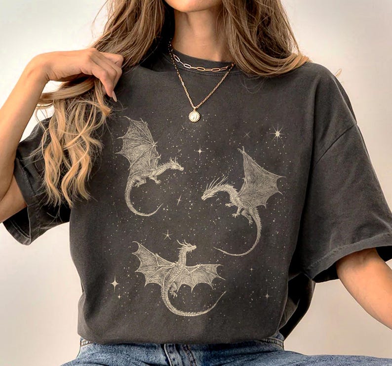 K&ouml;nnte beinhalten: Dunkelgraues T-Shirt mit drei detaillierten Drachenillustrationen mit ausgebreiteten Fl&uuml;geln vor einem Sternenhintergrund. Die Drachen sind in einer hellen Farbe gehalten, die einen Kontrast zur Farbe des Shirts bildet. Das Shirt hat eine lockere Passform.