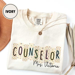 May include: Ivory-colored t-shirt with the word "COUNSELOR" in a floral design, outlined in black, green, and pink. The name "Mrs. Victoria" is written below in a script font. A gold necklace with a pendant is draped across the shirt.