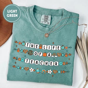 May include: A light green t-shirt with the text "THE LIFE OF A TEACHER" in white block letters. The shirt features colorful graphics of a globe, apple, rainbow, and other school-themed icons. A silver necklace with a pendant is on the shirt.