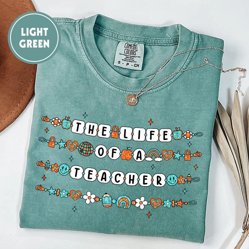 May include: A light green t-shirt with the text "THE LIFE OF A TEACHER" in white block letters. The shirt features colorful graphics of a globe, apple, rainbow, and other school-themed icons. A silver necklace with a pendant is on the shirt.
