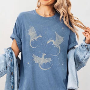 K&ouml;nnte beinhalten: Blaues T-Shirt mit einem Himmelsdesign, das drei detaillierte Drachenillustrationen zeigt. Die Drachen haben ausgebreitete Fl&uuml;gel und sind von sternartigen Akzenten umgeben. Das Shirt hat eine lockere Passform und gerollte &Auml;rmel.